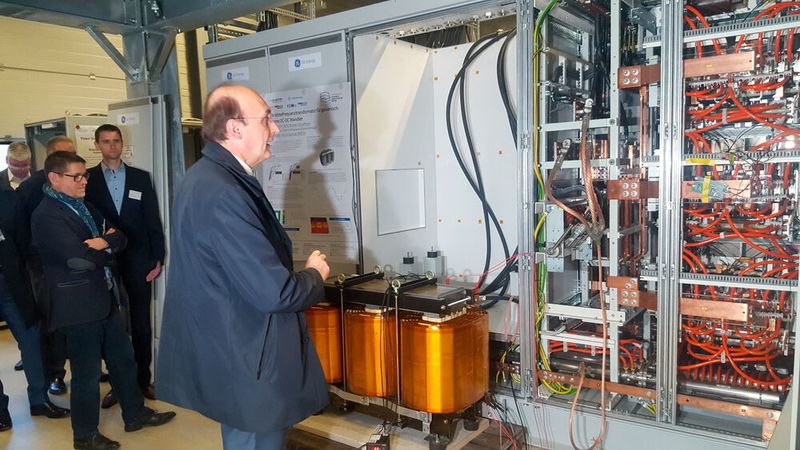 Prof. Rik De Doncker erklärt einer Delegation bei der Eröffnung das dreiphasige Dual-Active-Bridge(DAB)-Prinzip, nachdem der aufgebaute Gleichspannungswandler arbeitet und das einen bidirektionalen, flexibel steuerbaren Leistungsfluss ermöglicht und gleichzeitig eine galvanische Trennung bietet, womit die Anforderungen zukünftiger Gleichspannungsnetze erfüllt werden.
 (Energieagentur NRW)