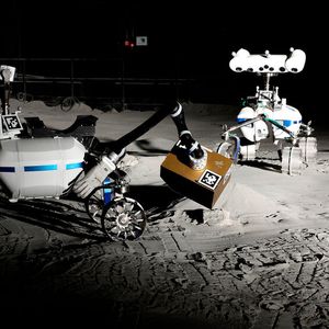 Die beiden Rover LRU2 (links) und LRU1 sind in der LUNA-Halle unterwegs. LRU2 richtet mit seinem Roboterarm das LIBS (Laser-Induced Breakdown Spectroscopy) aus, das sich in der goldfarbenen Box befindet. LIBS verwendet einen gepulsten Laser, um zum Beispiel einen Stein zu analysieren. Forschende haben in der LUNA-Halle Messwerkzeuge und Roboter getestet, die Wassereis auf dem Mond finden können.(Bild:  Roboter-Teamwork_ LRU1 und LRU2 arbeiten mit LIBS.jfif /DLR / CC BY 3.0)