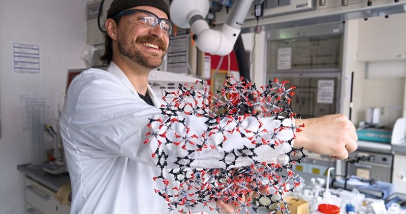 Kalle Mertin demonstriert die poröse Struktur von CAU-10-H anhand eines Modells: Dass sein Arm hindurchpasst, veranschaulicht die durchgehenden, röhrenartigen Poren, in denen das Material Wassermoleküle aufnimmt und wieder freisetzt. (Bild:  Christina Anders, Uni Kiel)