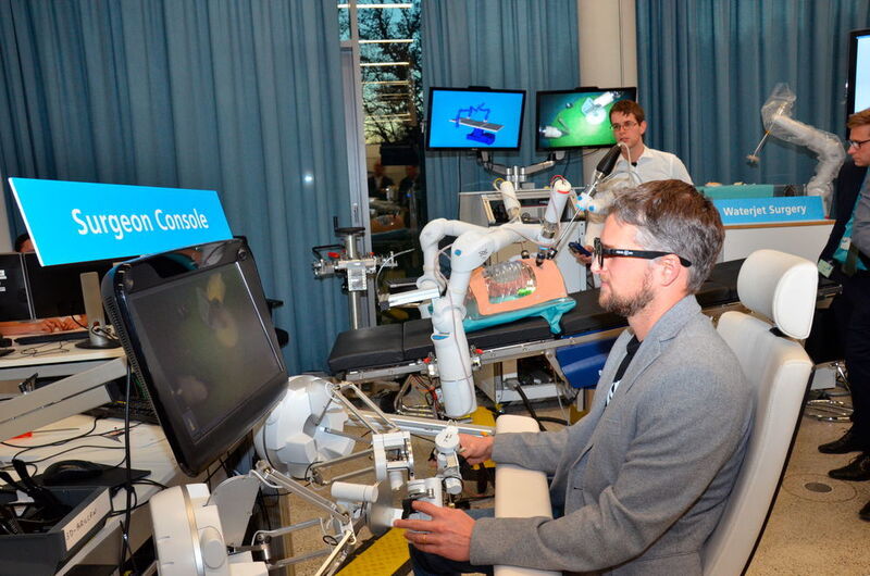 Chirurgenkonsole zur telepräsenten Ansteuerung von chirurgischen Instrumente im Inneren eines Patienten, im Hintergrund OP-Tische mit den Roboterarmen, die die Instrumente führen. (Forum MedTech Pharma e.V. / DLR)