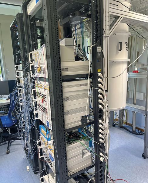 In der Realität sieht ein laufender Quantencomputer mit supraleitendem Quantenchip wenig aufregend aus, wie das Foto eines Systems von Peak Quantum in Garching bei München zeigt. Das komplexe Innenleben steckt in einer isolierten Hülle, die für die Kühlung auf eine Temperatur notwendig ist, die knapp über dem absoluten Nullpunkt liegt. Links daneben ist das Quantum Computing Control System von Zurich Instruments zu sehen, einem Tochterunternehmen von Rohde & Schwarz. (Bild: Vogel IT-Medien)