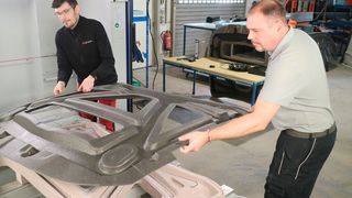 Mitarbeiter von Germa Composite legen zur Nachbearbeitung eine große Innenverstärkungsschale aus Faserverbundkunststoff für eine Fahrzeug-Fronthaube auf die Fräsvorrichtung, die bereits in der Gantry-5-Achs-Fräsanlage von HG Grimme Systech auf dem Vakuumspanntisch liegt.  (Bild: HG Grimme Systech / K. Rüsberg)