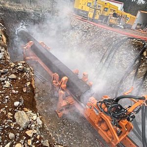 Die Druckluft aus den Atlas-Copco-Kompressoren (im Hintergrund) treibt den Schlagkolben des Imlochhammers im Inneren des Hammerbohrgeräts (groß im Bild) an, wodurch der Bohrkopf der Maschine auf die Bohrlochsohle schlägt. Durch die Schläge tritt am Kopf Luft aus, die zugleich die Sohle reinigt und das gelöste Bohrgut über den Ringraum der Maschine nach hinten und zurück an die Erdoberfläche spült.  (Bild: Petzold)