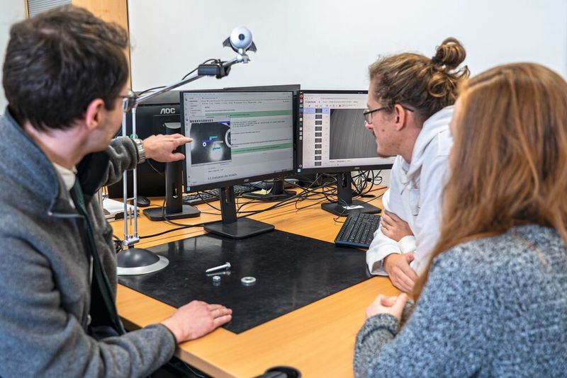 Die Webcam fotografiert die Schraube, Mutter und Unterlegscheibe auf der schwarzen Matte. Der Trainer (links) zeigt den beiden Azubis, wie sie die Kleinteile für die Software richtig labeln.  (Bild: Julia Thurner-Irmler, Universität Augsburg)