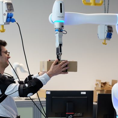 Forscher Federica Masiero nimmt ein Päckchen entgegen, dessen Gewicht der Roboter vorher bereits an das Exoskelett übermittelt hat. (Bild: Andreas Schmitz / TUM)