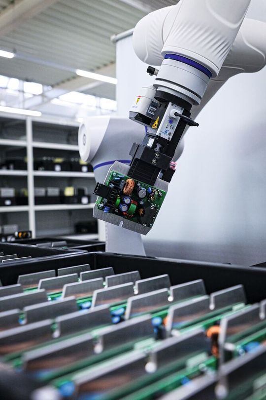 Handwerk trifft Automatisierung: Ein Cobot handhabt eine bestückte Baugruppe für eine Stromversorgung. Trotz des manufakturellen Charakters bei Spezialwandlern investiert das Unternehmen gezielt in moderne Fertigungstechniken, um die Prozesse im Stammhaus sowohl reproduzierbar als auch kosteneffizient zu gestalten.(Bild:  MTM Power)