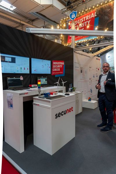 Am Stand von Secunet konnten Interessierte einem simulierten Angriff auf die Leitstelle eines Kraftwerks beiwohnen. Die Attacke richtet sich auf das OT-Netz der Windräder, die mit dem Produkt Secunet Edge gesichert sind. Die Angriffserkennung erfolgt über Secunet Monitor KRITIS, was in einem Dashboard auf Monitoren visualisiert wird. Gleichzeitig gibt eine angeschlossene Signallampe optische Hinweise auf die jeweilige Bedrohungslage. Zusätzlich hierzu erhält man über die Kommunikationsapp Stashcat Messenger eine Meldung auf das Smartphone, die einen ausführbaren Befehl zur direkten Reaktion auf den Angriff enthält. Über diesen kann man direkt aus der App reagieren und den Angriff isolieren. (Bild: Vogel Communications Group)
