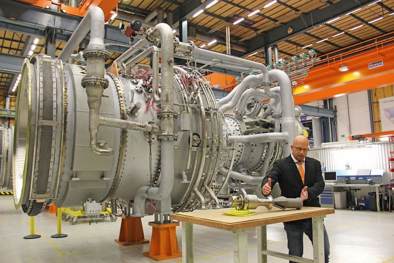 Auf dem Tisch im Vordergrund zeigt Andreas Graichen, Leiter des Teams für additive Fertigung, den Teil des Hauptantriebs, der gedruckt wurde. Im Hintergrund die Gasturbine, in der 28 solcher Antriebe verbaut sind. (Käfer)