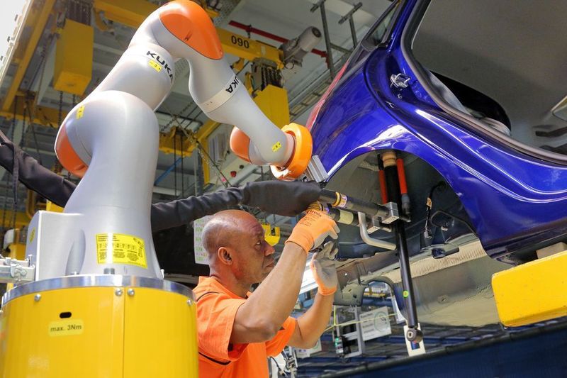 Zusammenarbeit von Mensch mit Maschine in der Ford-Fiesta-Fertigung: Ausgestattet mit Schrauben und Stoßdämpfer fährt der Leichtbauroboter automatisch in Richtung des Radhauses der Fahrzeug-Karosserie. Dort kontrolliert der Mitarbeiter noch einmal die Position, geführt über einen Haltegriff am Roboter, und gibt das Signal zum Schrauben. (Ford-Werke)