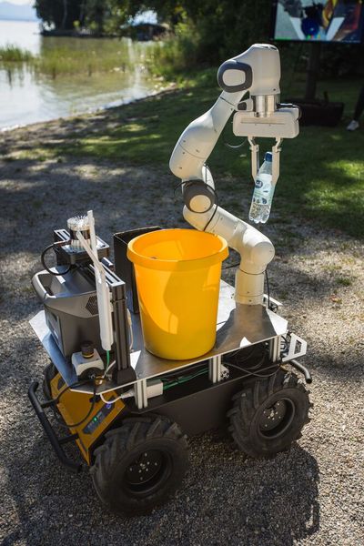 Der gelbe Landroboter sammelt mit seinem weißen Greifarm eine im Kies liegende Flasche auf und lässt diese in dem kleinen Eimer auf der Ladefläche verschwinden. (Bild: Hyundai)