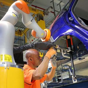Zusammenarbeit von Mensch mit Maschine in der Ford-Fiesta-Fertigung: Ausgestattet mit Schrauben und Stoßdämpfer fährt der Leichtbauroboter automatisch in Richtung des Radhauses der Fahrzeug-Karosserie. Dort kontrolliert der Mitarbeiter noch einmal die Position, geführt über einen Haltegriff am Roboter, und gibt das Signal zum Schrauben.