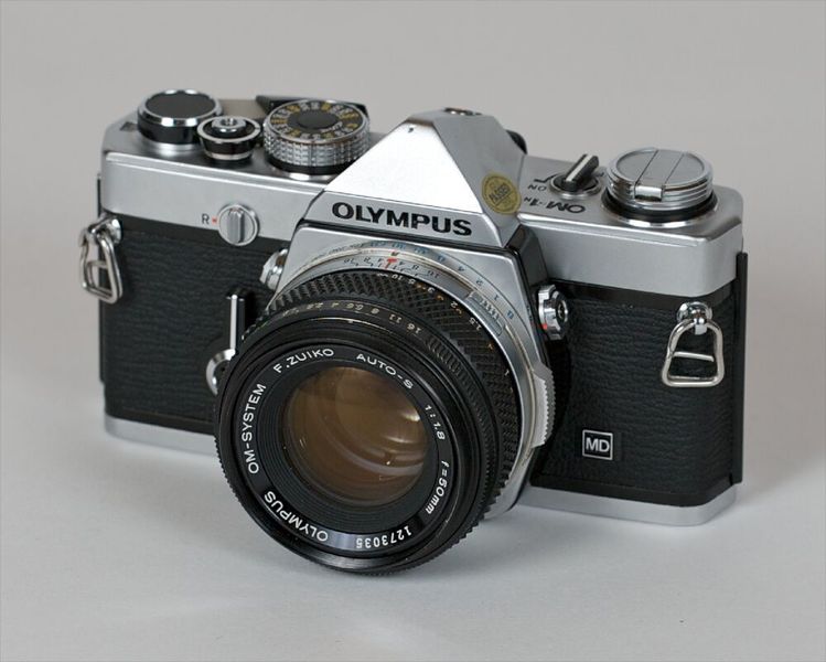 August Arnold brachte mit seinem Chefkonstrukteur E. Kästner die Spiegelreflexkamera zur Serienreife. Die 1938 patentierte Erfindung von Arnold bezieht sich nicht auf das einer Spiegelreflex-Filmkamera zugrundeliegende Prinzip, sondern auf die für die serienmäßige Herstellung der Kamera wesentlichen Komponenten: einen umlaufenden Blendenkörper aus Pressspiegelglas mit Versteifungsrippen sowie einer tellerförmigen Vertiefung des Pressglasblendenkörpers in seinem Mittelteil. (Olympus OM-1 / Benjamin Nagel / CC BY-SA 2.0)