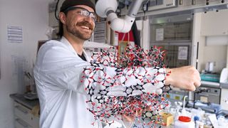 Kalle Mertin demonstriert die poröse Struktur von CAU-10-H anhand eines Modells: Dass sein Arm hindurchpasst, veranschaulicht die durchgehenden, röhrenartigen Poren, in denen das Material Wassermoleküle aufnimmt und wieder freisetzt.  (Bild: Christina Anders, Uni Kiel)