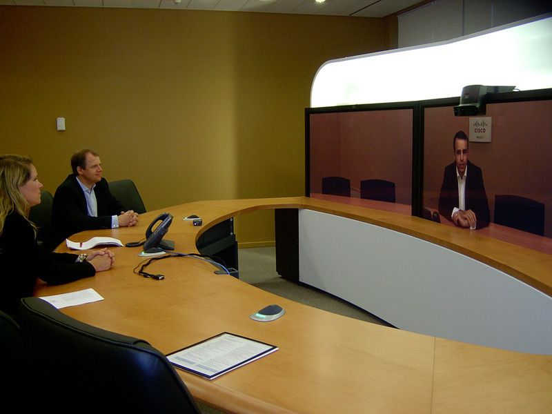 Abbildung 9: Das Video-Conferencing-System „Telepresence“ von Cisco findet sich ebenfalls im CBC. Christiph Plus, der technische Leiter des CBC, und Ernst Engelmann, der Projektverantwortliche, demonstrieren wie es wirkt und funktioniert. Plur sitzt in Eschborn und Engelmann in Neufahrn. Langt Engelmann mit seinem Arm über den Bildausschnitt hinaus, erscheint sein Arm im daneben liegenden Fenster. (Archiv: Vogel Business Media)
