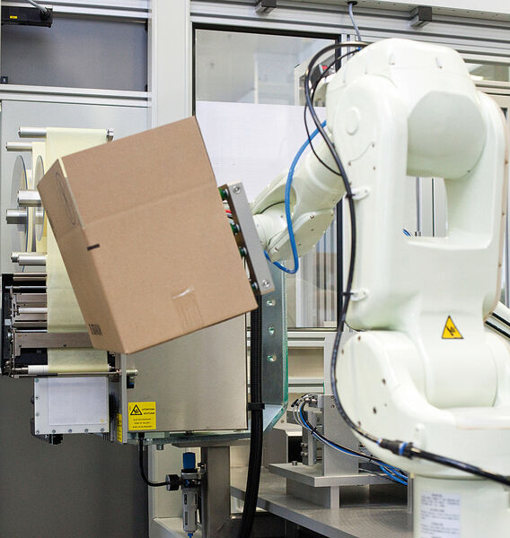 Automation: Ein Roboter führt den Karton vor den Etikettendruckspender. (Bild: Bluhm Systeme)