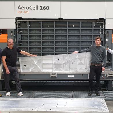 Proudly presenting the finished sample component in front of the AeroCell 160 | 400 (from left): Jens Ilg (Business Development, Aerospace & Composites MAPAL), Alexander Follenweider (Component Manager Aerospace & Composites MAPAL), Stefan Diem (application engineer bavius) and Dominik Merz (Director Global Sales bavius). In the foreground is the fixture used for clamping. (Source: MAPAL Dr. Kress SE & Co. KG )