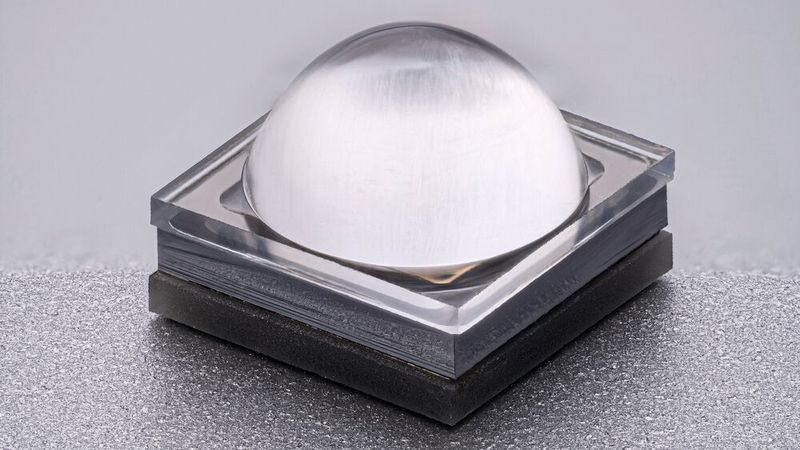 Fern-UVC-LED mit Linse und Reflektor: Nahaufnahme einer quadratischen LED mit transparenter, kuppelförmiger Linse auf dunkler Grundplatte. Das hermetisch geschlossene Gehäuse sorgt für höhere Zuverlässigkeit und Robustheit unter anspruchsvollen Umgebungsbedingungen.(Bild:  FBH_Schurian)