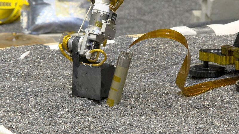 In einem JPL-Labor drückt ein Testmodell des Insight-Roboters mit seiner Schaufel auf den Boden in der Nähe des Testmodellss vom selbsthämmernden Marsmaulwurfs. DLR-Forscher haben berechnet, dass dem Maulwurf ein solches Drücken auf den Marsboden helfen kann, zu graben, da sich die Reibung des umgebenden Bodens erhöht. (NASA/JPL-Caltech)