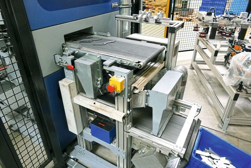 Oben das Einlaufband in die Entgratmaschine, unten das Auslaufband mit integriertem Platten-Entgratmagnetisierer und der nachpositionierten Transportkiste, die bei erreichtem Gewicht vom Roboter gegen eine leere Kiste ausgetauscht wird. Am Ende des Auslaufbandes werden Reststäube abgesaugt. Hinten rechts ist die Roboter-Werkzeugwechselstation erkennbar.  (Kuhn)