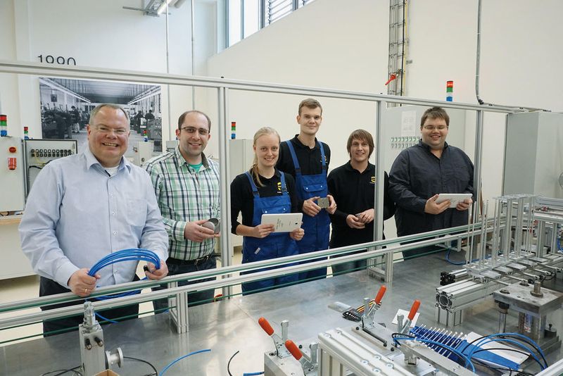 Der Ausbildungsleiter Nico Gottlieb (links), die Ausbilder Artur Maurer (zweiter von links) und Jacob Knierim (rechts) und die duale Studentin Adina Brandt, Maschinenbau, der Auszubildende Rino Richter, Mechatroniker, und der Duale Student Pascal Haßebrock, Mechatronik, an der Ausbildungseinheit Industrie 4.0: Hier können Blechdosen per Smartphone individuell mit Schokoladentäfelchen bestückt werden. (Harting)
