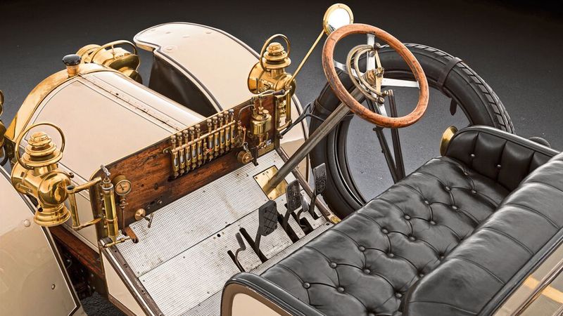 Die Bedienung eines Automobils kurz nach der Jahrhundertwende unterschied sich aber noch spürbar – Beispiel Pedale. So hatte der Simplex derer vier: Neben der Kupplung (links) befinden sich zwei Pedale für die zwei unabhängig voneinander arbeitenden Außenband-Bremsen am Getriebe. Der Handbremshebel (außen, rechts vom Fahrer) war für die hinteren Trommelbremsen zuständig. In der Mitte des „Armarturenbretts“ befindet sich die sogenannte „Schmiertrompete“ für die Ölversorgung der verschiedenen Reibstellen am Fahrzeug. (Bild:  Mercedes-Benz AG)