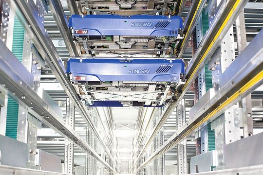 Herzstück der Logistikerweiterung 2014: Im vollautomatischen Shuttlelager fahren 375 Shuttles durch die 15 jeweils 60 m langen Regalgassen. Wenn die Artikelansprache es erfordert, wechseln die Shuttles (siehe Bild) über Shuttleheber die Lagerebenen und haben somit auf insgesamt 200.000 Behälter mit bis zu 800.000 Lagerplätzen Zugriff. Herzstück der Logistikerweiterung 2014: Im vollautomatischen Shuttlelager fahren 375 Shuttles durch die 15 jeweils 60 m langen Regalgassen. Wenn die Artikelansprache es erfordert, wechseln die Shuttles (siehe Bild) über Shuttleheber die Lagerebenen und haben somit auf insgesamt 200.000 Behälter mit bis zu 800.000 Lagerplätzen Zugriff.