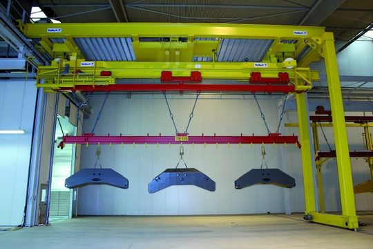 Bild 1: Am Beginn der Großteilelackieranlage übernimmt der Be- und Entlademanipulator die Transporteinheiten (rot) mit den eingehängten Werkstücken. Je nach Bedarf können die Gehänge mit Einzelteilen oder mehreren Werkstücken bis 20 t bestückt werden. Bild 1: Am Beginn der Großteilelackieranlage übernimmt der Be- und Entlademanipulator die Transporteinheiten (rot) mit den eingehängten Werkstücken. Je nach Bedarf können die Gehänge mit Einzelteilen oder mehreren Werkstücken bis 20 t bestückt werden.
