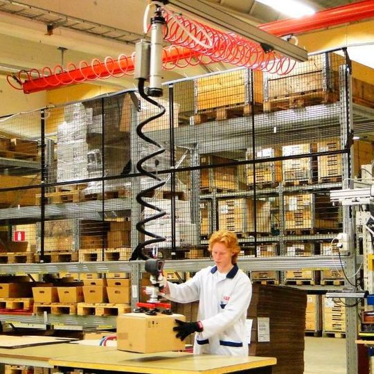 Ein Mitarbeiter hebt ergonomisch und mühelos einen Karton mit einem „Basic“-Allround-Seilbalancer von 3i. Ein Mitarbeiter hebt ergonomisch und mühelos einen Karton mit einem „Basic“-Allround-Seilbalancer von 3i.
