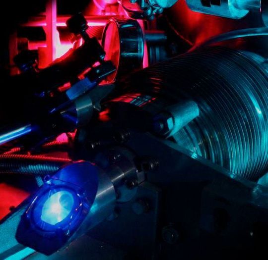 Dieses Foto zeigt den Vakuumapparat, der zur Messung der 2S-4P-Übergangsfrequenz in atomarem Wasserstoff genutzt wurde. Das violette Leuchten im Hintergrund stammt aus der Mikrowellenentladung, die Wasserstoff-Moleküle in Wasserstoff-Atome dissoziiert. Das blaue Licht im Vordergrund ist Fluoreszenz vom ultravioletten Laser, der die Atome in den 2S-Zustand anregt. Das türkisfarbene Leuchten ist Streulicht von dem Lasersystem, das zur Frequenzmessung des 2S-4P-Übergangs dient. Dieses Foto zeigt den Vakuumapparat, der zur Messung der 2S-4P-Übergangsfrequenz in atomarem Wasserstoff genutzt wurde. Das violette Leuchten im Hintergrund stammt aus der Mikrowellenentladung, die Wasserstoff-Moleküle in Wasserstoff-Atome dissoziiert. Das blaue Licht im Vordergrund ist Fluoreszenz vom ultravioletten Laser, der die Atome in den 2S-Zustand anregt. Das türkisfarbene Leuchten ist Streulicht von dem Lasersystem, das zur Frequenzmessung des 2S-4P-Übergangs dient.