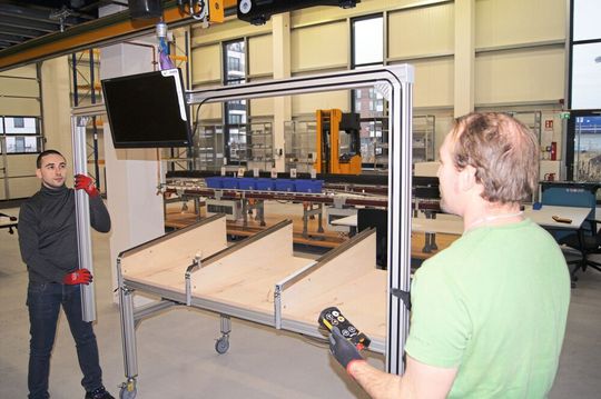 Die Hängebahnanlage unterhalb der Stahlbühne im Versuchsfeld bietet Unterstützung bei leichten Handling-Aufgaben, hier zum Beispiel bei der Montage eines Kommissioniertisches.