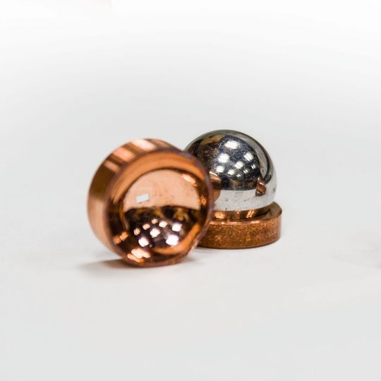 Copper mirrors direct and focus microwave photons in electrons on liquid helium system. The focusing mirror (on the left) is made by pressing a hard stainless steel ball (also shown in the picture) into a soft piece of copper. The flat mirror (on the right) shows two concentric circular electrodes (aka a Corbino pair) that are used to measure electron conductivity. Copper mirrors direct and focus microwave photons in electrons on liquid helium system. The focusing mirror (on the left) is made by pressing a hard stainless steel ball (also shown in the picture) into a soft piece of copper. The flat mirror (on the right) shows two concentric circular electrodes (aka a Corbino pair) that are used to measure electron conductivity.