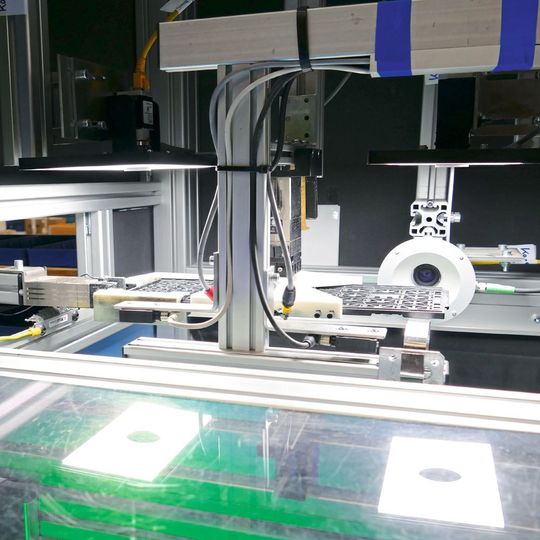 Zwei Kameras nehmen Bilder der Trays von oben, eine von der Seite auf. In der Spiegelung sind die beiden oberen Beleuchtungen erkennbar, links übernimmt der Roboterarm die geprüften Trays. Zwei Kameras nehmen Bilder der Trays von oben, eine von der Seite auf. In der Spiegelung sind die beiden oberen Beleuchtungen erkennbar, links übernimmt der Roboterarm die geprüften Trays.