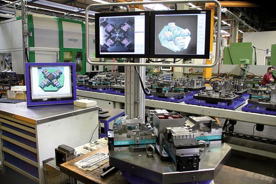 Visuelles Assistenzsystem für einen Arbeitsplatz zur Bestückung von Wechselpaletten. Im Vordergrund ist die drehbar gelagerte Bauteilaufnahme, im Hintergrund die Visualisierung auf Bildschirmen mit Kameraüberlagerung und CAD-Darstellung zu sehen. (Bild: Kolbus)