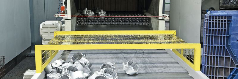 Aluminiumdruckgussteile am Auslauf einer Drahtgurt-Strahlanlage. Mit diesem Anlagentyp lassen sich Massenteile kontinuierlich mit Aluminium strahlen.
