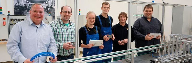 Der Ausbildungsleiter Nico Gottlieb (links), die Ausbilder Artur Maurer (zweiter von links) und Jacob Knierim (rechts) und die die Duale Studentin Adina Brandt (Maschinenbau), der Auszubildende Rino Richter (Mechatroniker) und der Duale Student Pascal Haßebrock (Mechatronik) an der „Ausbildungseinheit Industrie 4.0“: Blechdosen können per Smartphone individuell mit Schokoladentäfelchen bestückt werden.