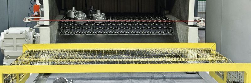 Die-cast aluminium parts at the outlet of a wire belt blasting machine. With this type of machine, mass-produced parts can be continuously blasted with aluminium.