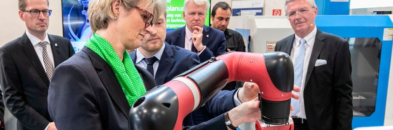 Lukas Nikelowski (Fraunhofer-IML) hilft Ministerin Karliczek bei der Bedienung eines Roboterarms. Im Hintergrund (v.l.): Prof. Dr. Georg Rosenfeld, Ullrich Sierau, Georgios Katsimitsoulias und Prof. Dr. Michael ten Hompel.