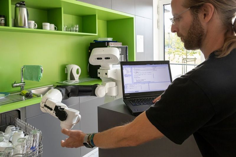 Roboter Tiago reicht in einer Küche des DFKI in Osnabrück einem Wissenschaftler eine Tasse aus der Spülmaschine. Die Maschinen sollen vorübergehende Objektverbindungen erkennen lernen.