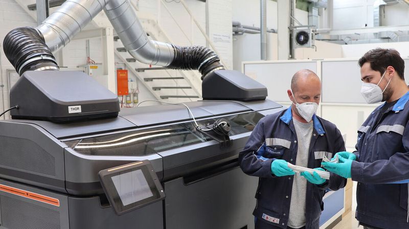 Volkswagen-Mitarbeiter prüfen die Qualität von im Binder-Jetting-Verfahren hergestellten Bauteilen. Im Hintergrund der Prototyp des Druckers.