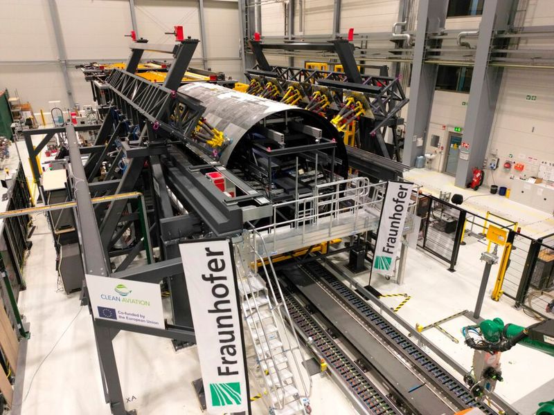 Montage-Forschungsplattform mit eingerüsteten thermoplastischen Rumpfschalen des „MFFD“ bei der Fraunhofer-Gesellschaft in Stade. Gut erkennbar sind die gelben Hexapod-Roboter zum Halten und hochpräzisen Einstellen von Form sowie Lage der Oberschale. (Bild: Fraunhofer IFAM)