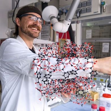 Kalle Mertin demonstriert die poröse Struktur von CAU-10-H anhand eines Modells: Dass sein Arm hindurchpasst, veranschaulicht die durchgehenden, röhrenartigen Poren, in denen das Material Wassermoleküle aufnimmt und wieder freisetzt.  (Bild: Christina Anders, Uni Kiel)