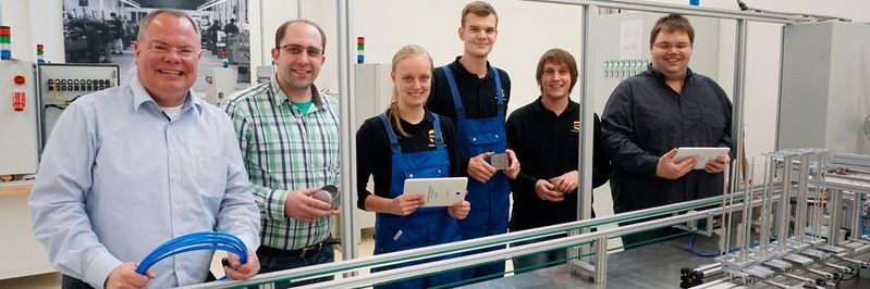 Nico Gottlieb (Ausbildungsleiter), Artur Maurer (Ausbilder), Adina Brandt (duale Studentin Maschinenbau), Rino Richter (Auszubildender zum Mechatroniker), Pascal Haßebrock (dualer Student Mechatronik) und Jacob Knierim (Ausbilder; von links nach rechts) an der „Ausbildungseinheit Industrie 4.0“: Blechdosen können per Smartphone individuell mit Schokoladentäfelchen bestückt werden.(Bild:  Harting)