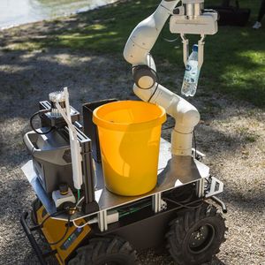 Der gelbe Landroboter sammelt mit seinem weißen Greifarm eine im Kies liegende Flasche auf und lässt diese in dem kleinen Eimer auf der Ladefläche verschwinden.(Bild:  Hyundai)