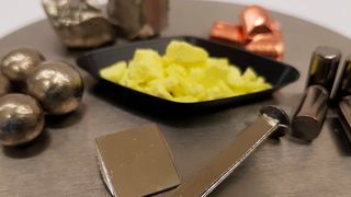 Amorphe Proben aus einer neuen Titan-Schwefel-Legierung, die drei junge Doktoranden der Universität des Saarlandes gemeinsam mit dem Technologiekonzern Heraeus entwickelt haben. Sie soll gleichzeitig leicht und fest sein (im Hintergrund, von links nach rechts, sind die Bestandteile der neuen Legierung zu sehen: Nickel, Zirkonium, Schwefel, Kupfer, Titan). (Oliver Gross/Universität des Saarlandes)