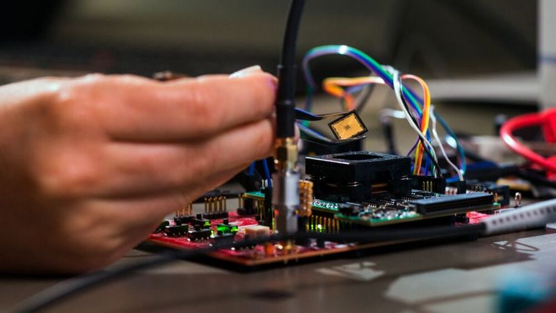 Tim Fritzmann von der Post-Quanten-Chip Forschung am Lehrstuhl Sicherheit in der Informationstechnik hält den PQC-Chip mit einer Pinzette, um diesen anschließend auf der Testplatine zu platzieren. Durch den Prozessorsockel (schwarzes Bauteil unter dem Chip) kann der Chip schnell ausgetauscht und eingebaut werden.(Bild:  Astrid Eckert / TUM)