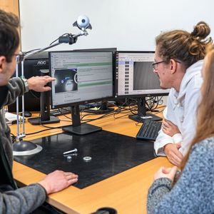 Die Webcam fotografiert die Schraube, Mutter und Unterlegscheibe auf der schwarzen Matte. Der Trainer (links) zeigt den beiden Azubis, wie sie die Kleinteile für die Software richtig labeln. (Bild:  Julia Thurner-Irmler, Universität Augsburg)