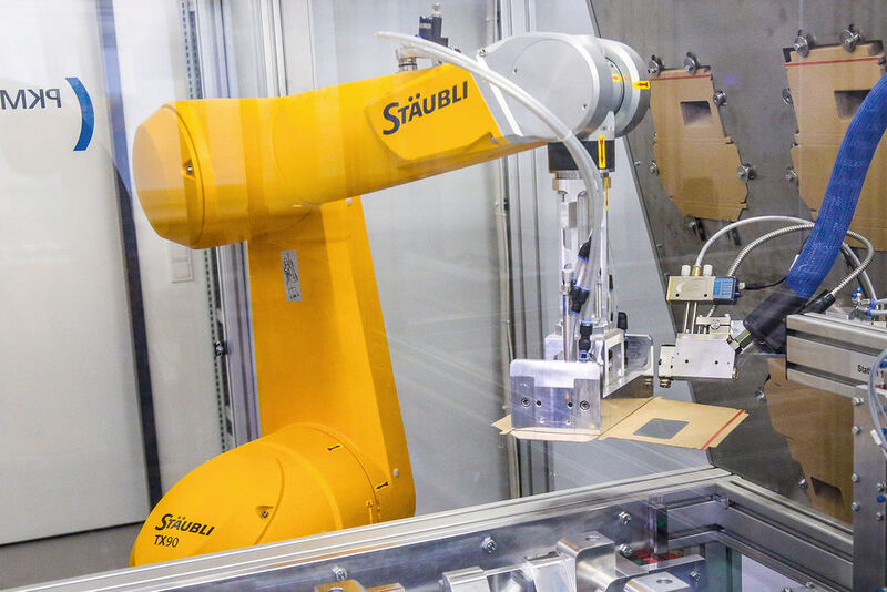 Per Roboterarm wird die benötigte Zuschnittgröße aus dem Vorrat entnommen, mit Heißkleber bestrichen und über eine Aufrichtschablone in den Transportwagen eingesetzt. (MPA NRW)